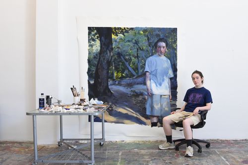 A photograph of artist Sian Costello sitting to the right of one of her paintings displayed in the upcoming show 'Hymn for Him'. The painting is a self-portrait of Costello standing in a forest scene. There is a table in the left of the photograph with various paints, brushes and similar implements.
